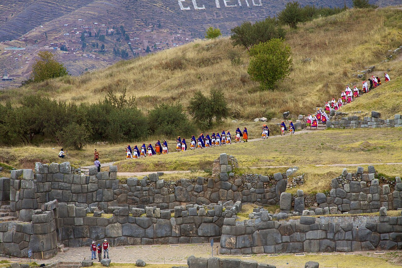 A long procession of costumed participants winding down a hillside past the stone walls of Sacsayhuaman during Inti Raymi