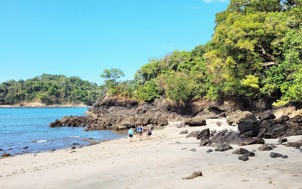 A pale sand beach backed by jungle with dark rocks in the foreground at Manuel Antonio