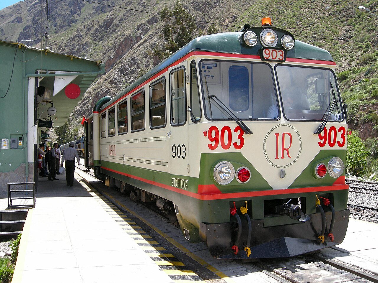 A green and cream Inca Rail railcar numbered 903 at a platform in a steep valley