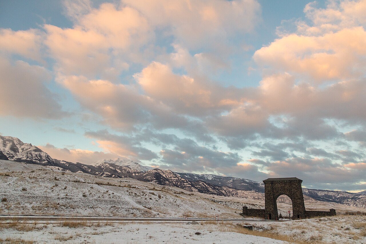 The stone Roosevelt Arch at Yellowstone's north entrance at sunrise, with snow-dusted hills and pink clouds behind