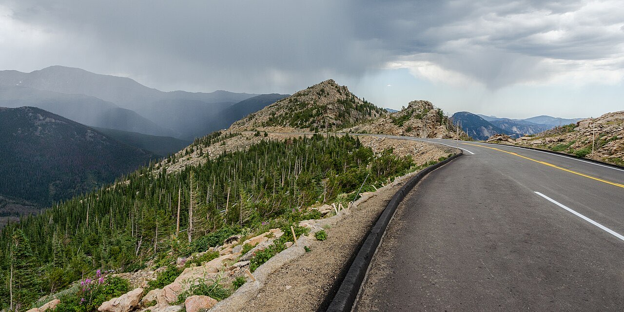 A paved two-lane road curving along a high alpine ridge in Rocky Mountain National Park with storm cloud over the peaks beyond