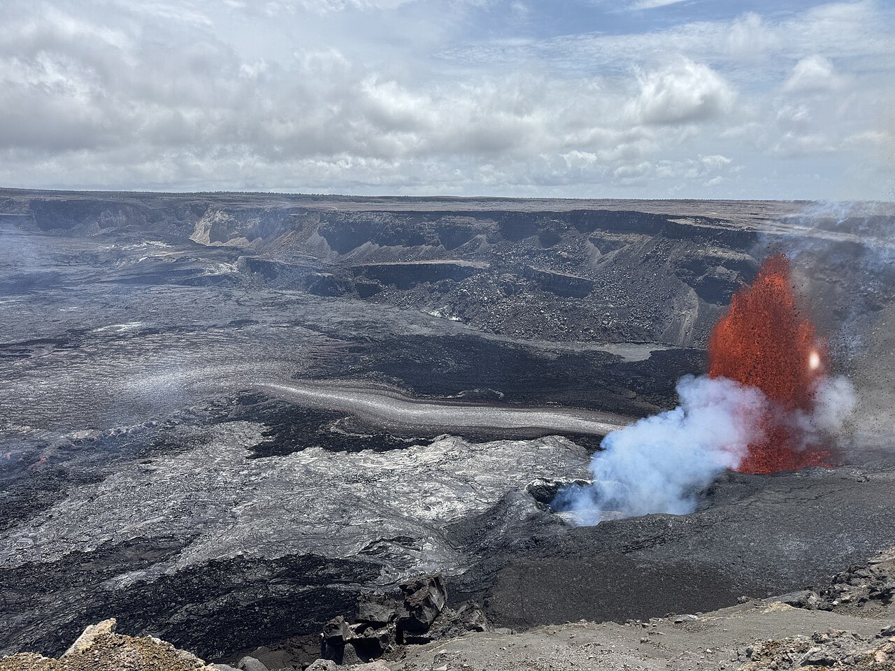 A lava fountain erupting from a vent inside the Halemaumau crater at Kilauea, with steam and dark crater floor around it